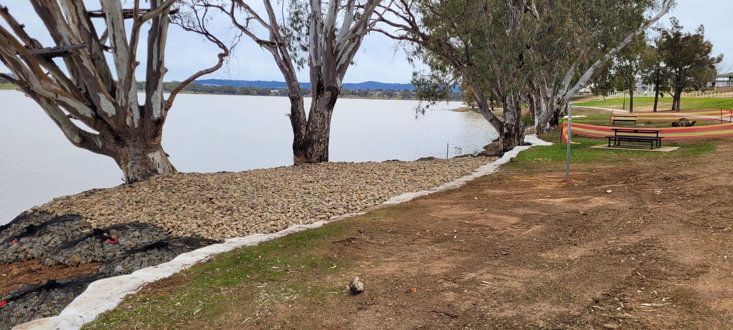 A lakefront area with a row of gum trees along the water's edge, where erosion control works are underway. The shoreline has been reinforced with rocks and mesh, and the ground is bordered by a white erosion control fabric. A picnic table sits on a concre
