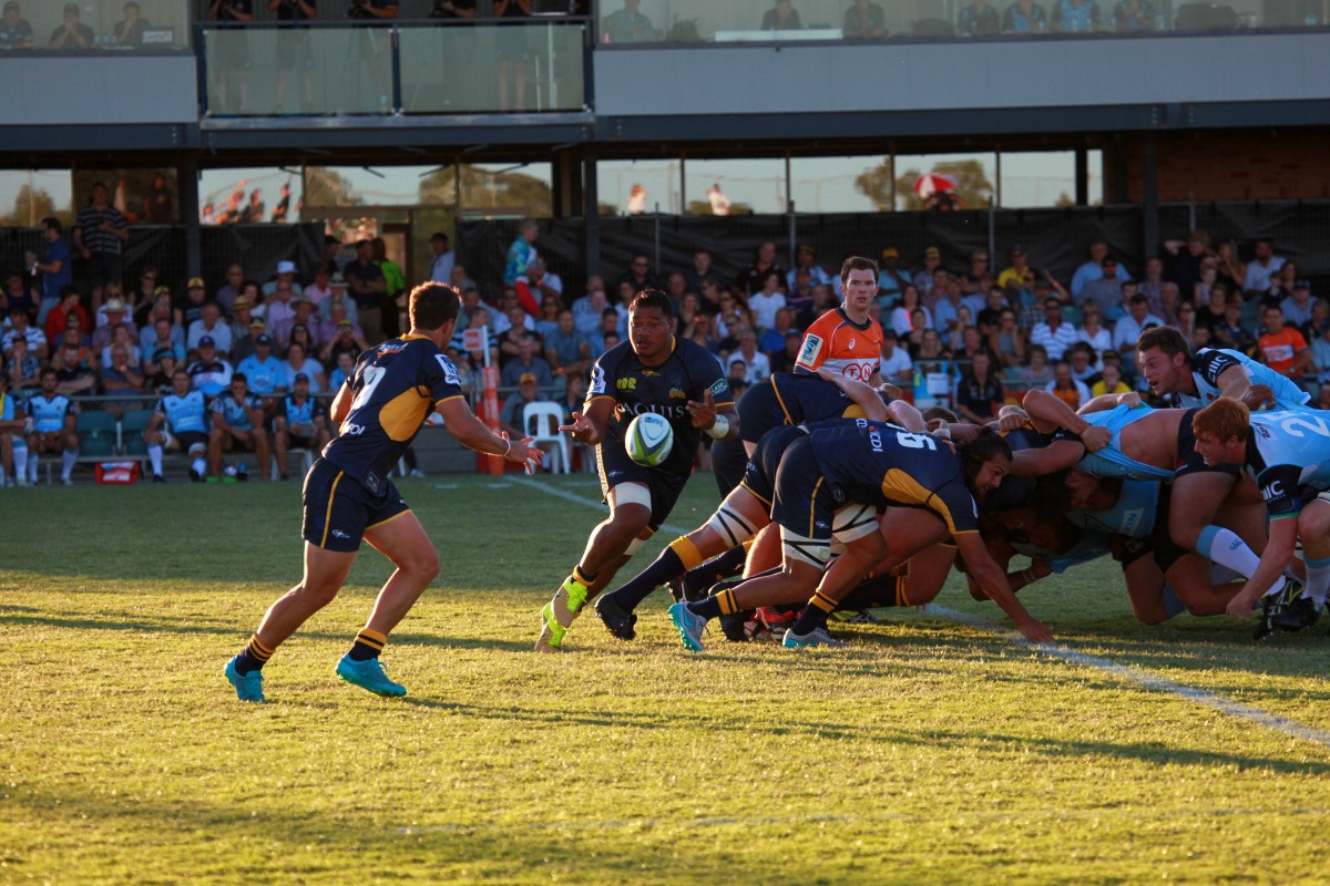 Rugby player passes the ball to another player, with other players still in a scrum.