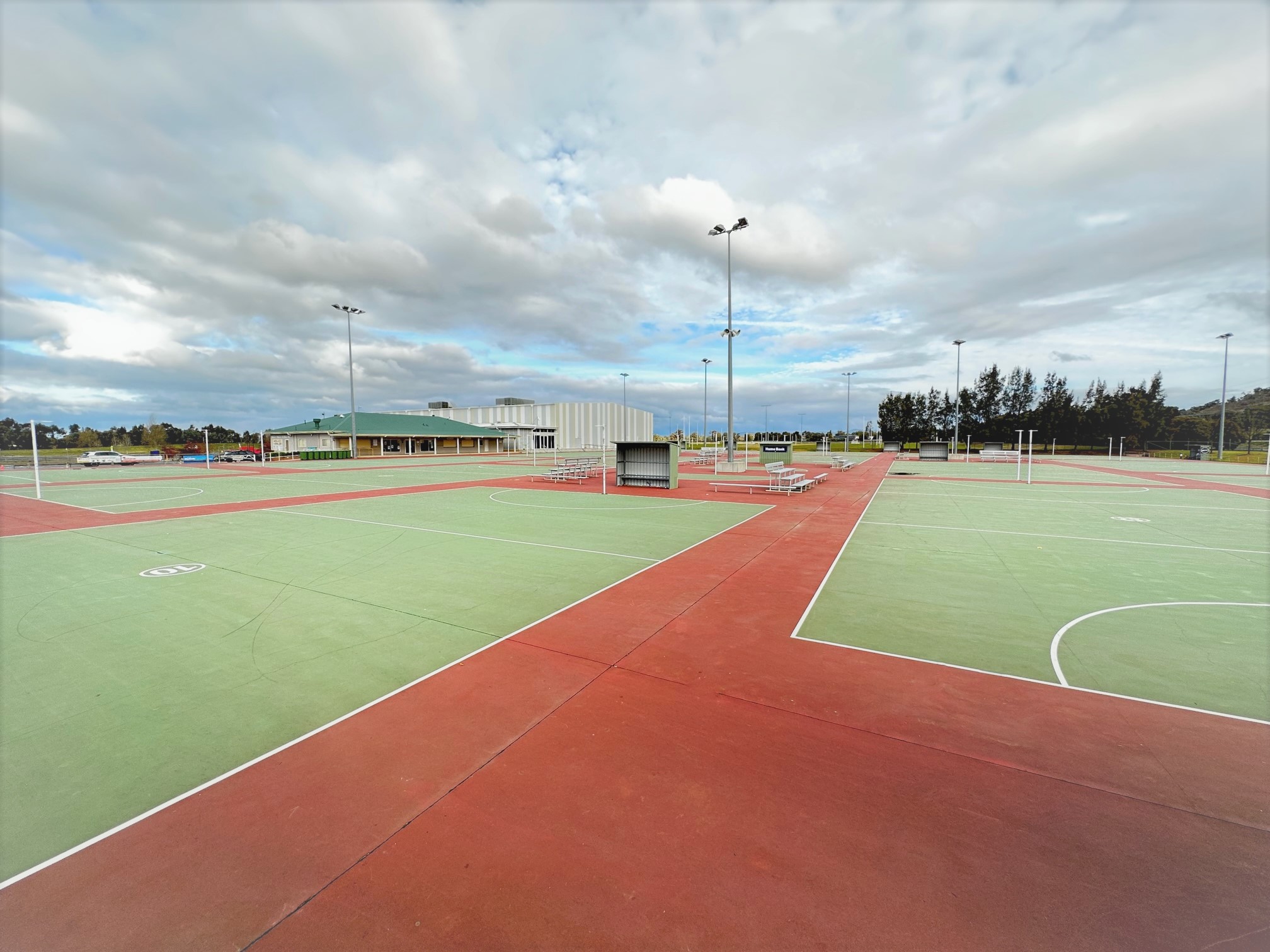 Netball courts in the foreground with club rooms and large stadium in the background, on a cloudy day.