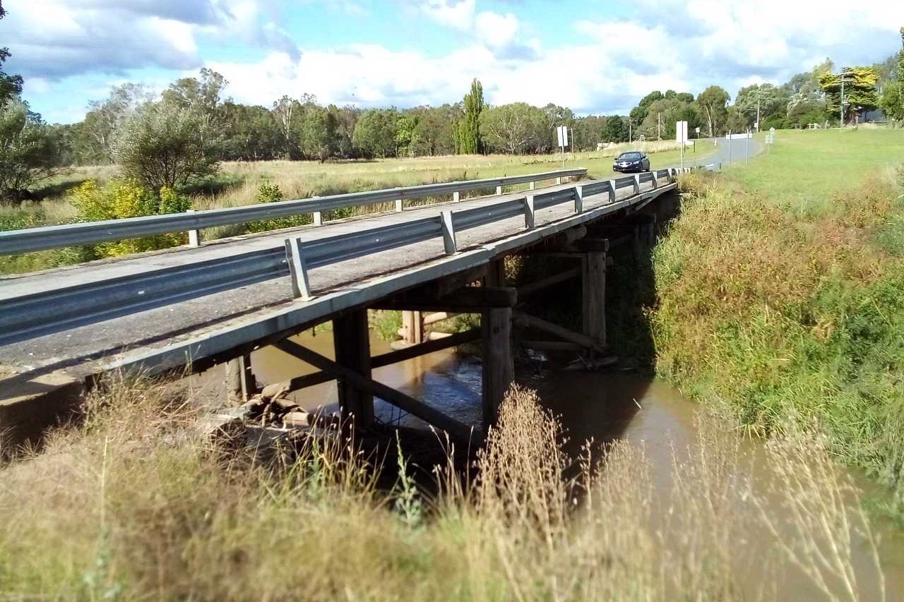 A wooden bridge over a creek with water in the creek, and a car and paddocks in the background.