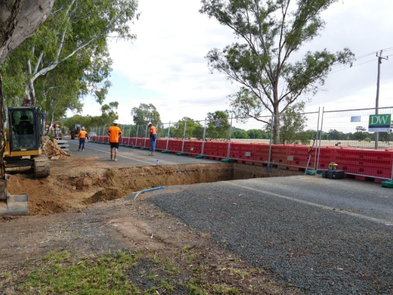 Three workers in orange hi-visibility shirts standing near a large, excavated trench on a road.