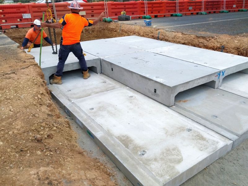 Two workers in orange hi-visibility shirts guide in concrete culverts into a large, excavated trench on a road.