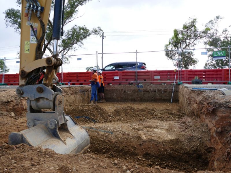 Earthmoving equipment removing soil under a road in the foreground, with workers, a mesh security fence, and a vehicle driving past in the background.