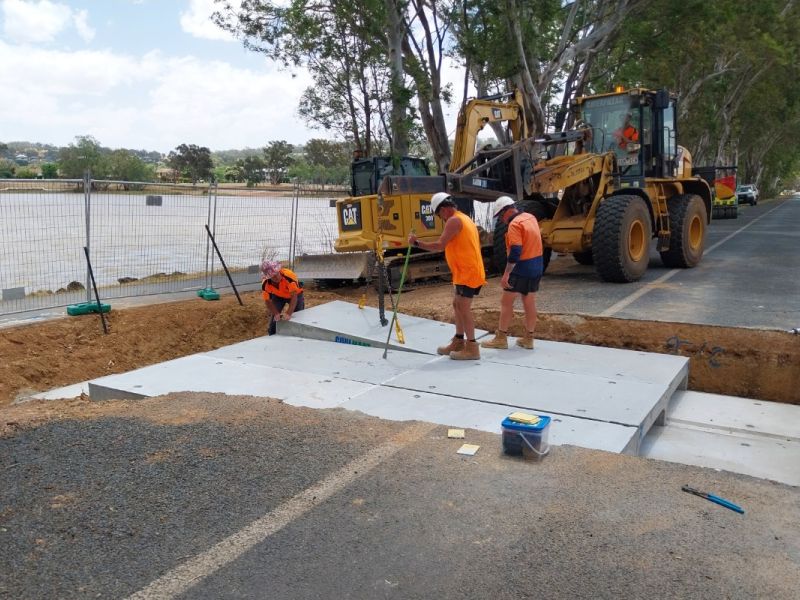 Three workers in orange hi-visibility shirts installing concrete culverts into a large, excavated trench on a road, with heavy earthmoving machinery in the background.