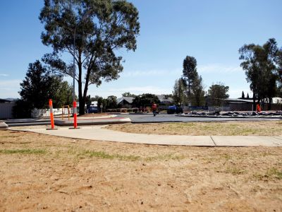 A man in an orange shirt walks across a fresh layer of asphalt on a roundabout under construction, with a concrete footpath in the foreground.