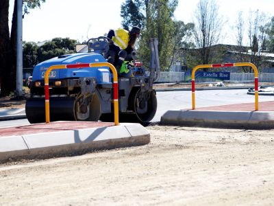 A man with a long beard and wearing a hi-vis shirt driving a mini-roller machine over a fresh layer of asphalt on a road.