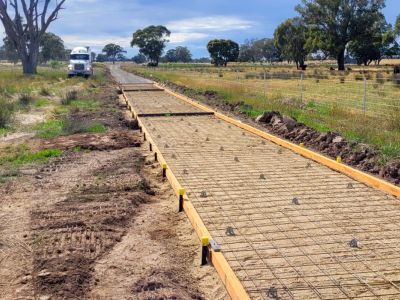 Form work for concrete pour on path through open grassed space, with cattle in a nearby paddock and a truck in the background.