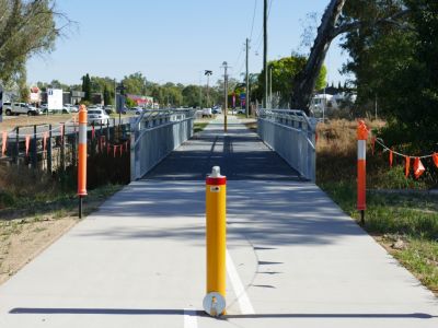 Yellow bollard in foreground on a concrete shared path with a steel footbridge in the background.