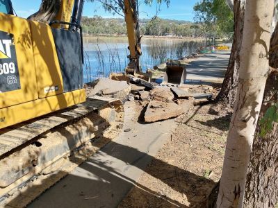A yellow earth moving machine scooping our soil and sections of asphalt foot and cycle path beside a lake.